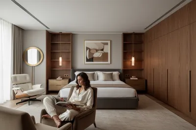 A woman sits in a chair reading in a luxurious bedroom. The room features a large bed, wooden cabinets, and a round mirror.