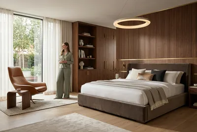 A woman stands in a luxurious bedroom with a large bed, wooden paneling, and a view of a garden.
