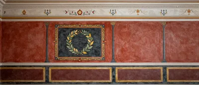 A symmetrical fresco of an ancient Roman interior wall. The wall is divided into three registers, with a central panel featuring a laurel wreath. The colors are predominantly red, black, gold, and green.