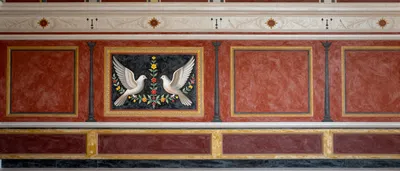 A photorealistic fresco of an ancient Roman wall with a central panel depicting two doves and floral garlands, flanked by red panels and decorative elements.
