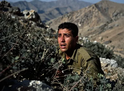 A soldier in military fatigues looks up with a look of fear, partially hidden behind mountain vegetation. Mountains are in the background.