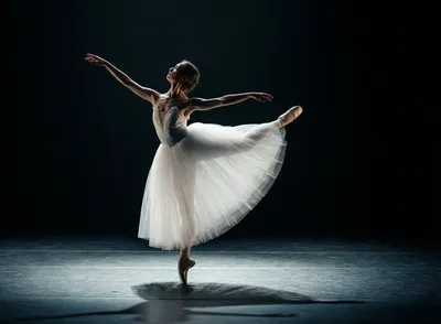 A ballerina in a white tutu performs an arabesque on a dark stage, lit by a spotlight.