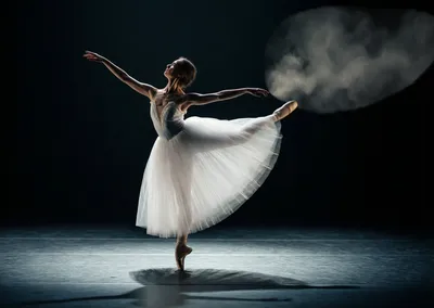 A ballerina in a white tutu balances en pointe, arms outstretched, under a spotlight.