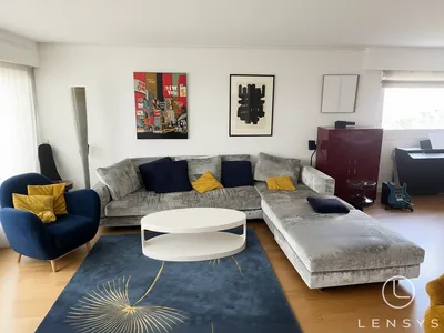 A bright, clean living room featuring a large grey L-shaped sectional sofa, a blue armchair, a white coffee table, and a blue rug with a gold botanical design.