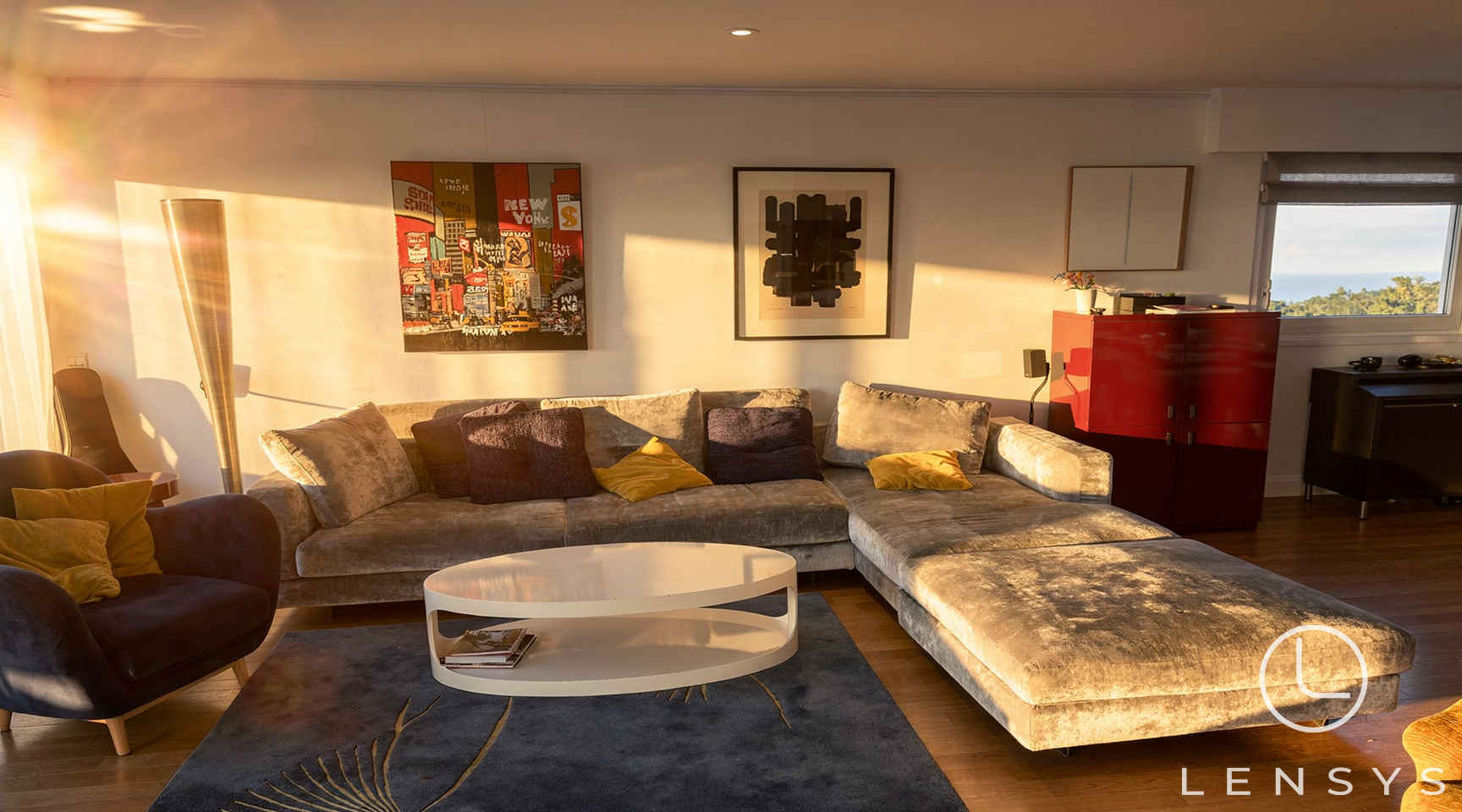 A modern living room bathed in warm sunlight, featuring a large sectional sofa, artwork, and a coffee table.