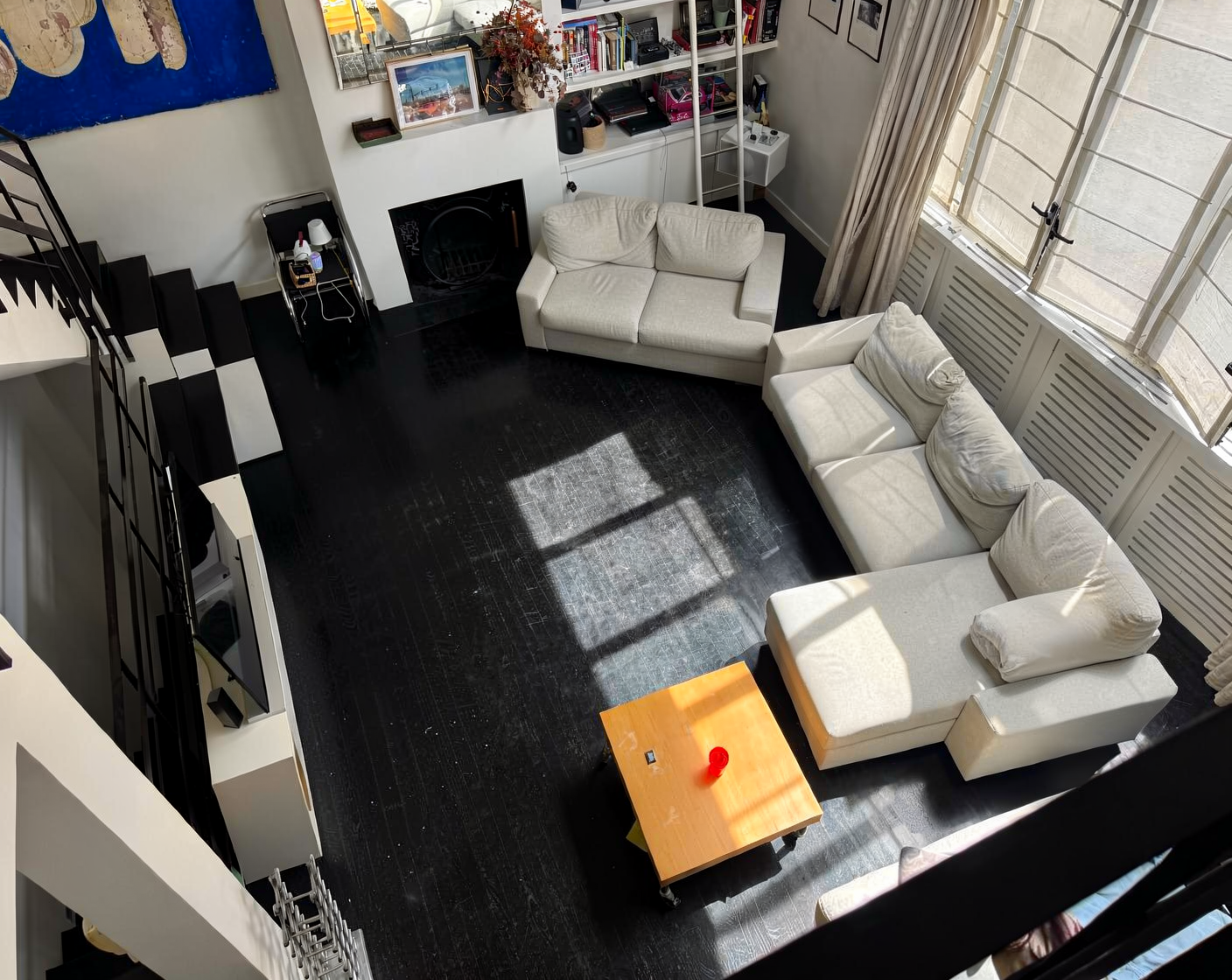 An overhead shot of a modern living room with a black wooden floor, white sofas, and large windows.