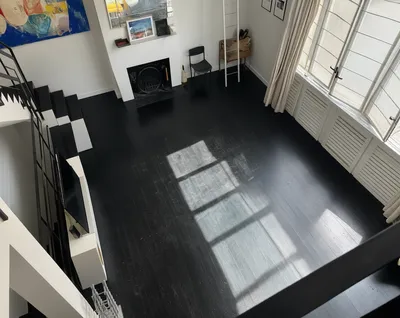 An empty, modern loft interior with dark wood floors, white walls, a black and white staircase, and large windows with natural light.