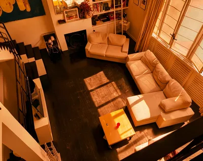 An overhead view of a sunlit living room with dark wooden floors, beige sofas, a wooden coffee table, and a staircase with black and white steps.