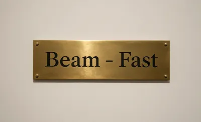 A rectangular brass plaque with the words "Beam - Fast" engraved in black serif font, mounted on a light off-white wall.