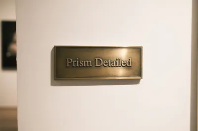 A rectangular brass museum placard with the words "Prism Detailed" engraved in serif typeface, mounted on a warm off-white wall.