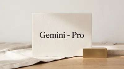 A cream-colored debossed letterpress card with "Gemini - Pro" in black serif text, standing on a wooden surface with a linen cloth and a brass detail.