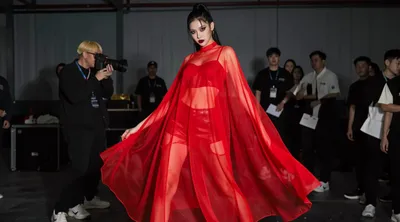 A model in a sheer red outfit poses confidently, with a photographer and crew visible in the background.